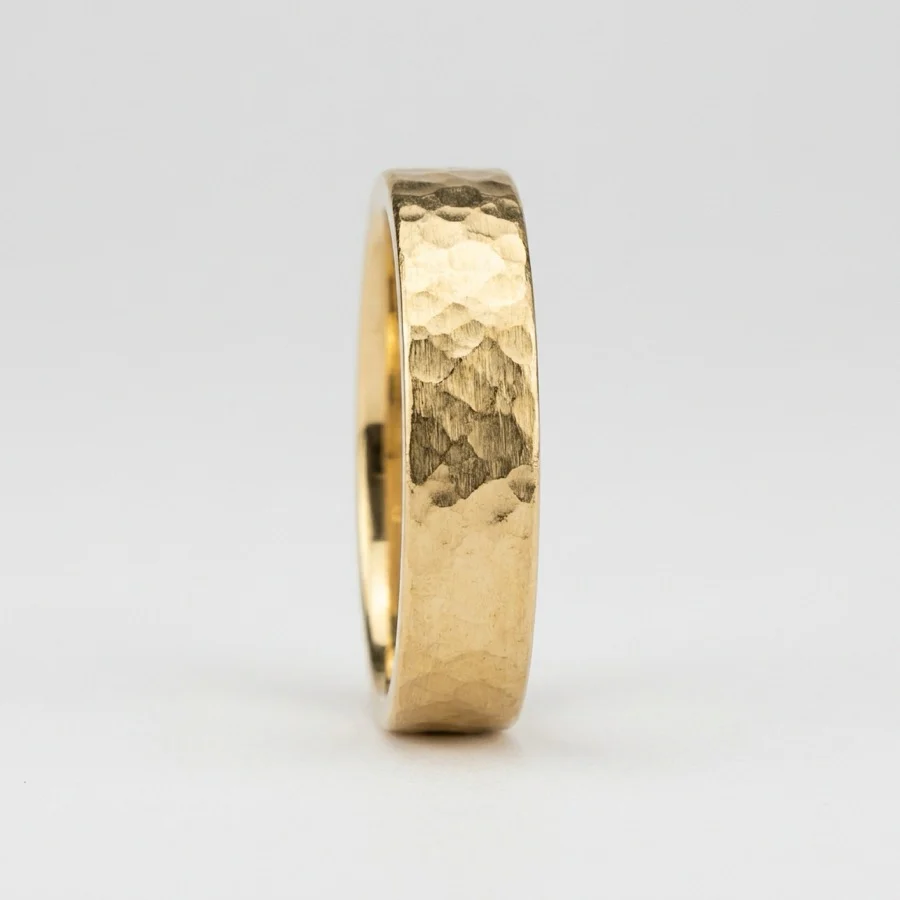 A product photograph of a wide hammered gold band ring standing upright on a plain light grey background, the beaten texture clearly visible