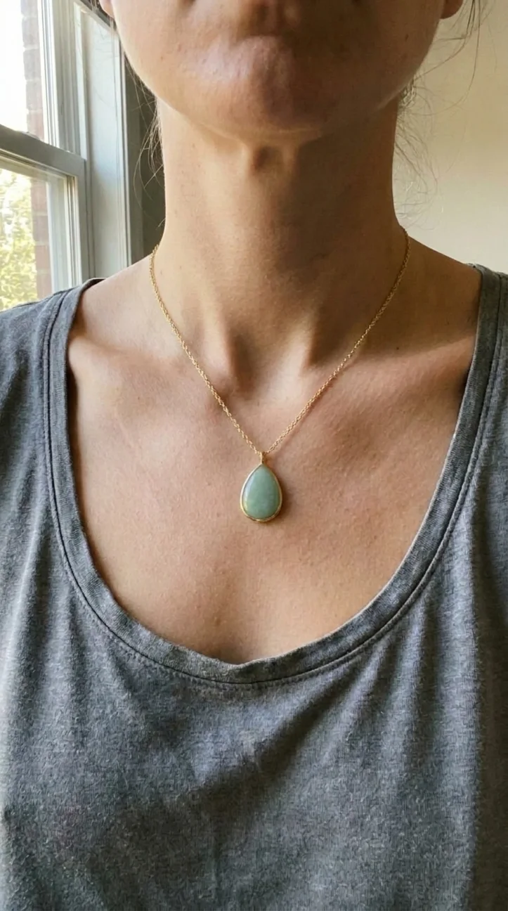 AI generated worn shot: a pale green teardrop pendant on a fine gold chain, worn at a woman's neckline over a grey vest top